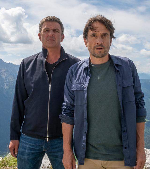 "Der Bergdoktor: Aus Mut gemacht": Dr. Martin Gruber (Hans Sigl) und Tim Hauser (Oliver Mommsen) stehen erhöht auf einem Berg und schauen an der Kamera vorbei angespannt in die Ferne. Hinter ihnen Bergpanorama.