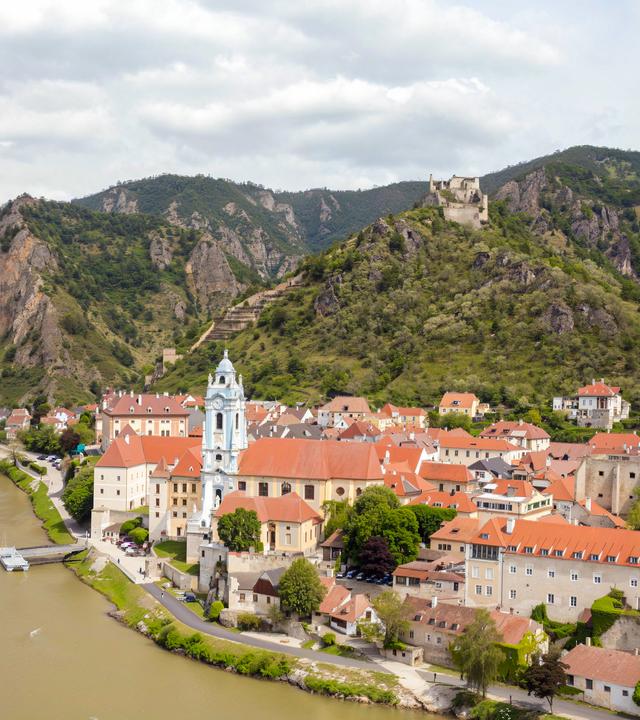 Blick auf Dürnstein in der Wachau mit der Donau im Vordergrund und den umliegenden Bergen im Hintergrund.