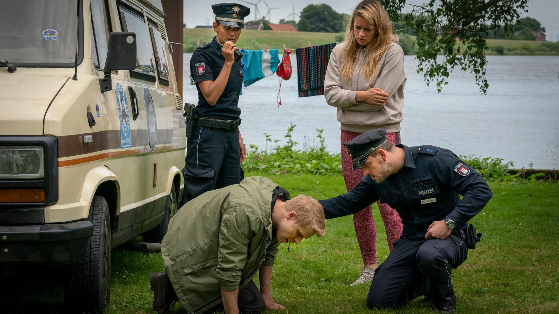 "Notruf Hafenkante - Ausgebremst": Die Polizistin Pinar Aslan (Aybi Era) steht neben einem Van, der auf einer Wiese an einem Gewässer steht. Sie spricht in ihr Funkgerät, das sie in ihrer rechten Hand hält. Neben ihr steht Linn (Chiara von Galli). Sie schaut besorgt zu Ben (Karl Schaper), der vor ihr auf dem Rasen kauert. Der Polizist Kris Freiberg (Marc Barthel) kniet vor ihm. Er hat fürsorglich seine rechte Hand auf Bens linken Schulter liegen.