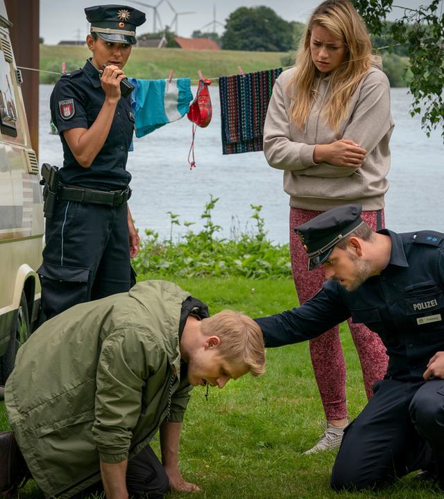 "Notruf Hafenkante - Ausgebremst": Die Polizistin Pinar Aslan (Aybi Era) steht neben einem Van, der auf einer Wiese an einem Gewässer steht. Sie spricht in ihr Funkgerät, das sie in ihrer rechten Hand hält. Neben ihr steht Linn (Chiara von Galli). Sie schaut besorgt zu Ben (Karl Schaper), der vor ihr auf dem Rasen kauert. Der Polizist Kris Freiberg (Marc Barthel) kniet vor ihm. Er hat fürsorglich seine rechte Hand auf Bens linken Schulter liegen.