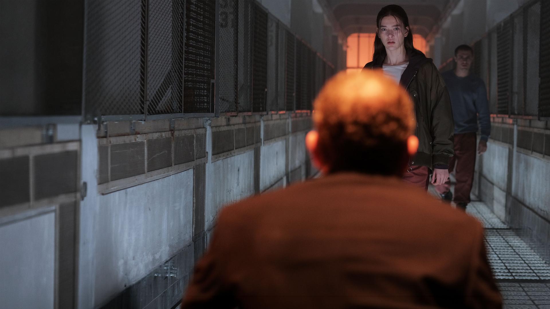 "Fallen - Ausgeflogen": Luce (Jessica Alexander) steht dem Outcast (Rod Hill) mit ernstem Blick gegenüber. Hinter ihr steht Daniel (Gijs Blom) in einem düsteren Gang mit Gitterwänden zu beiden Seiten.