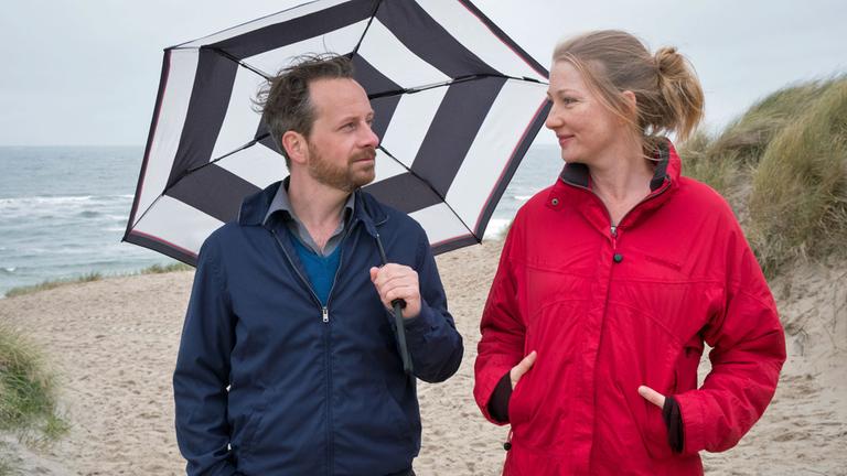 "Ausgerechnet Sylt": Kevin (Fabian Busch) und Bente (Katja Studt) spazieren gemeinsam am Strand entlang. Er hat einen Regenschirm über sich aufgespannt und schaut Bente lächelnd an, die seinen Blick erwidert. Im Hintergrund ist das Meer zu sehen.