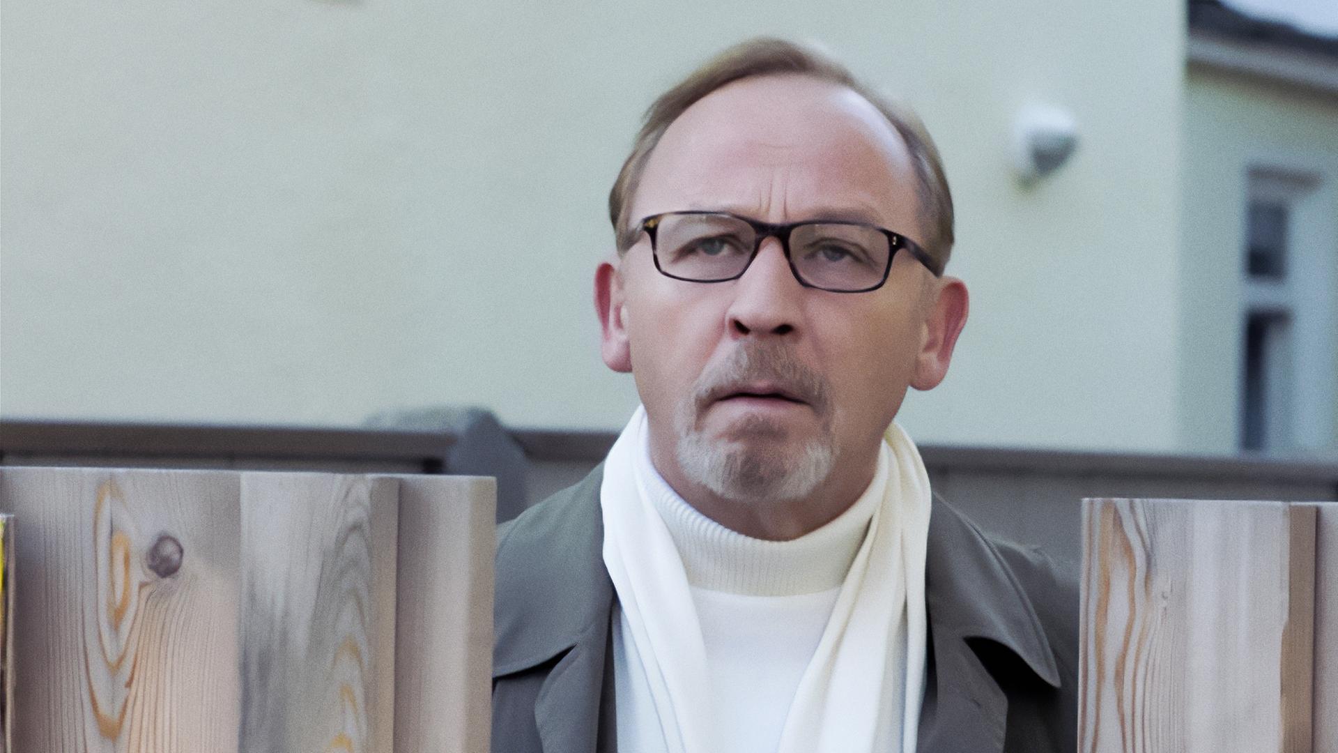 Ludwig Schaller (Alexander Held), der einen grauen Manzel und einen weißen Rollkragenpulli trägt, steht in einem Garten, vor ihm ein brusthoher Lattenzaun, über den er drüber späht. 