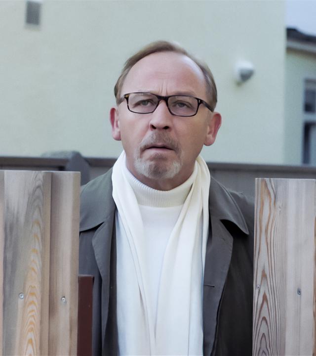 Ludwig Schaller (Alexander Held), der einen grauen Manzel und einen weißen Rollkragenpulli trägt, steht in einem Garten, vor ihm ein brusthoher Lattenzaun, über den er drüber späht. 