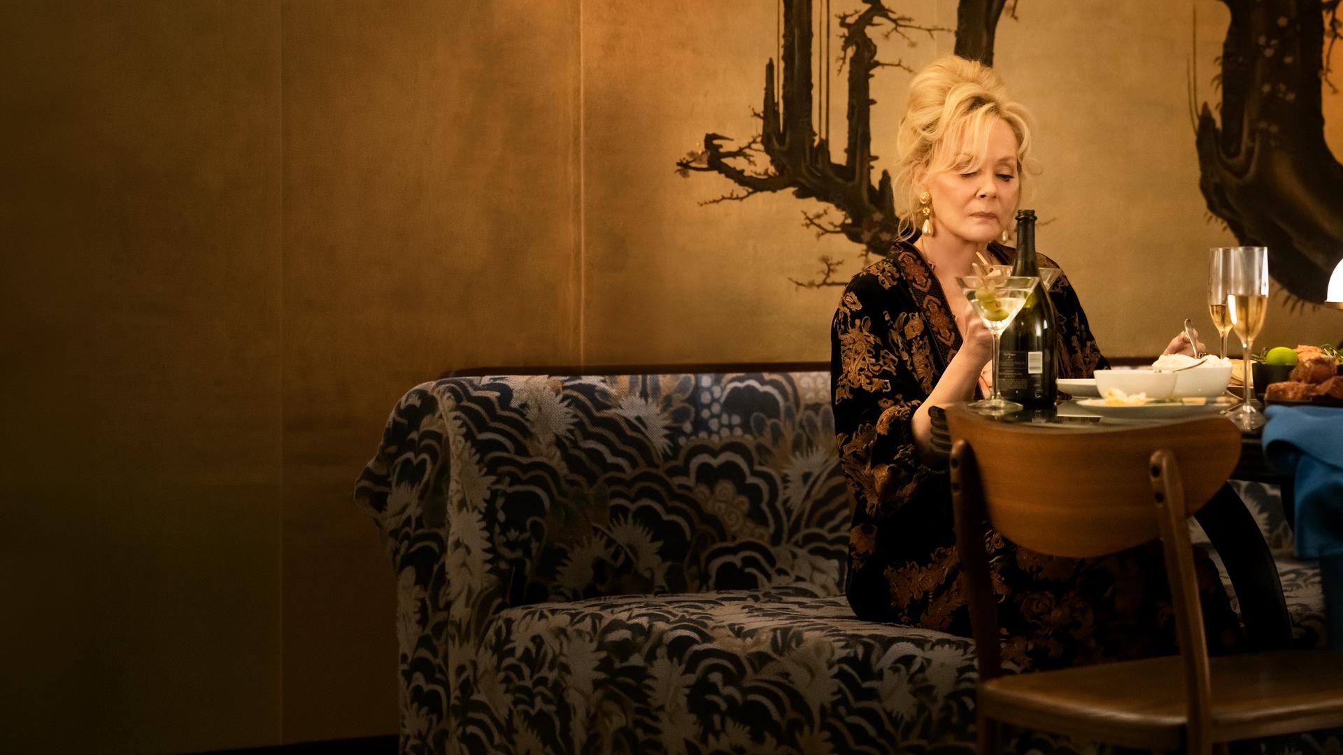 "Hacks - Ausschweifungen, hier und da": Deborah (Jean Smart) sitzt alleine an einem elegant gedeckten Tisch und hält ein Martini-Glas in der Hand, auf das sie nachdenklich blickt. Hinter ihr befindet sich eine kunstvoll gestaltete Wand mit einem baumartigen Muster.
