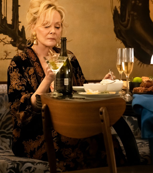 "Hacks - Ausschweifungen, hier und da": Deborah (Jean Smart) sitzt alleine an einem elegant gedeckten Tisch und hält ein Martini-Glas in der Hand, auf das sie nachdenklich blickt. Hinter ihr befindet sich eine kunstvoll gestaltete Wand mit einem baumartigen Muster.