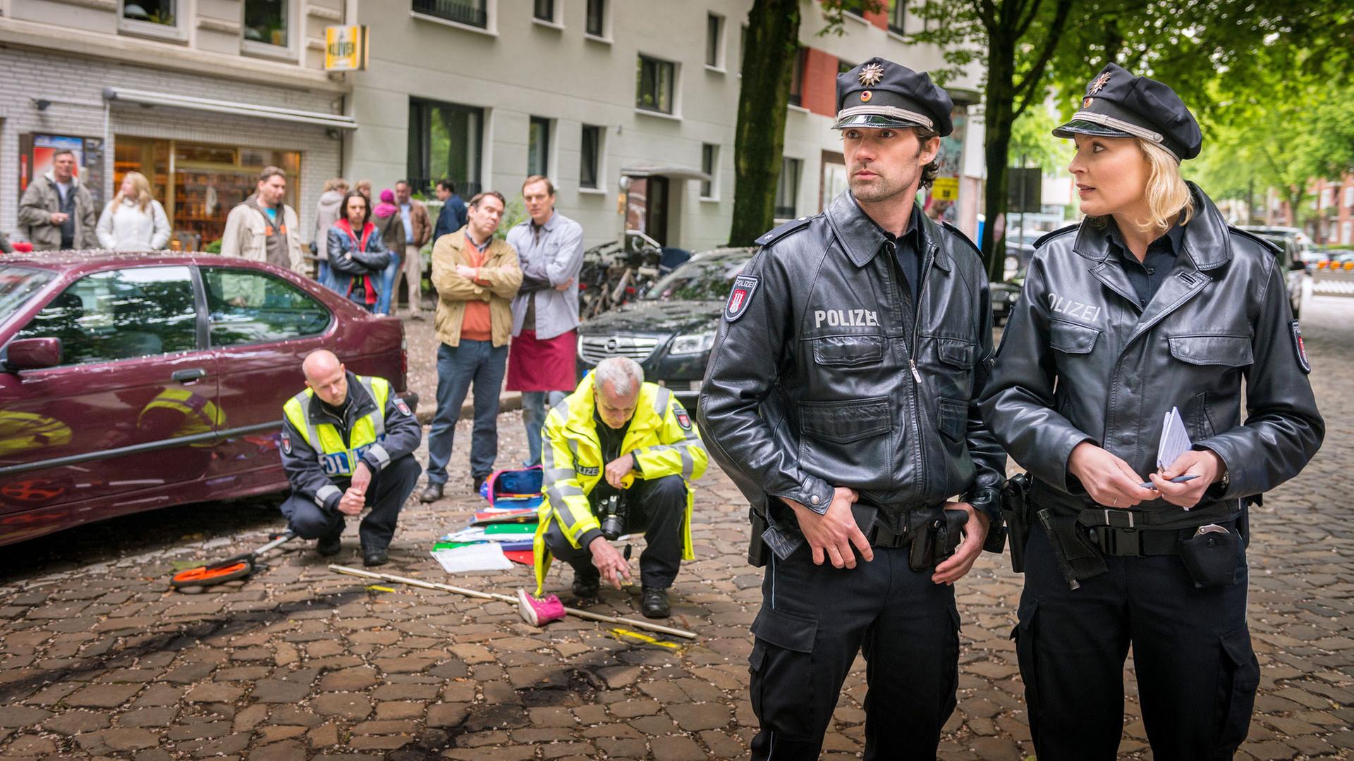 "Notruf Hafenkante - Außer Kontrolle": Am Straßenrand stehen Passanten, die zwei Polizeibeamte beobachten, die auf dem Boden der Straße eine Bremsspur vermessen und einzeichnen. Im Vordergrund die Polizeibeamten Mattes Seeler (Matthias Schloo) und Melanie Hansen (Sanna Englund), die versuchen, sich ein Bild des Unfallhergangs zu machen.