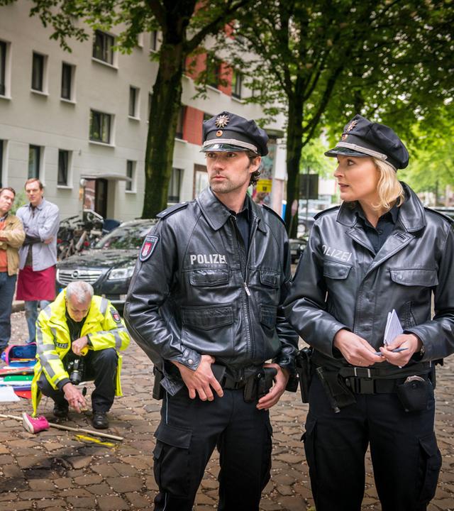 "Notruf Hafenkante - Außer Kontrolle": Am Straßenrand stehen Passanten, die zwei Polizeibeamte beobachten, die auf dem Boden der Straße eine Bremsspur vermessen und einzeichnen. Im Vordergrund die Polizeibeamten Mattes Seeler (Matthias Schloo) und Melanie Hansen (Sanna Englund), die versuchen, sich ein Bild des Unfallhergangs zu machen.