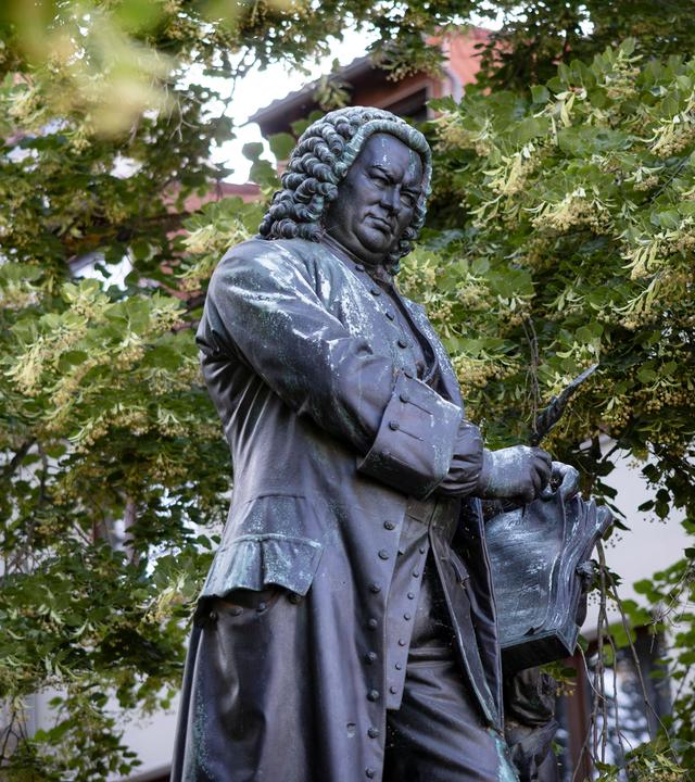 Statue des Komponisten Johann Sebastian Bach - man sieht die Skulptur eines Mannes in historischem Gehrock vor grünen Bäumen vor einem Gebäude.