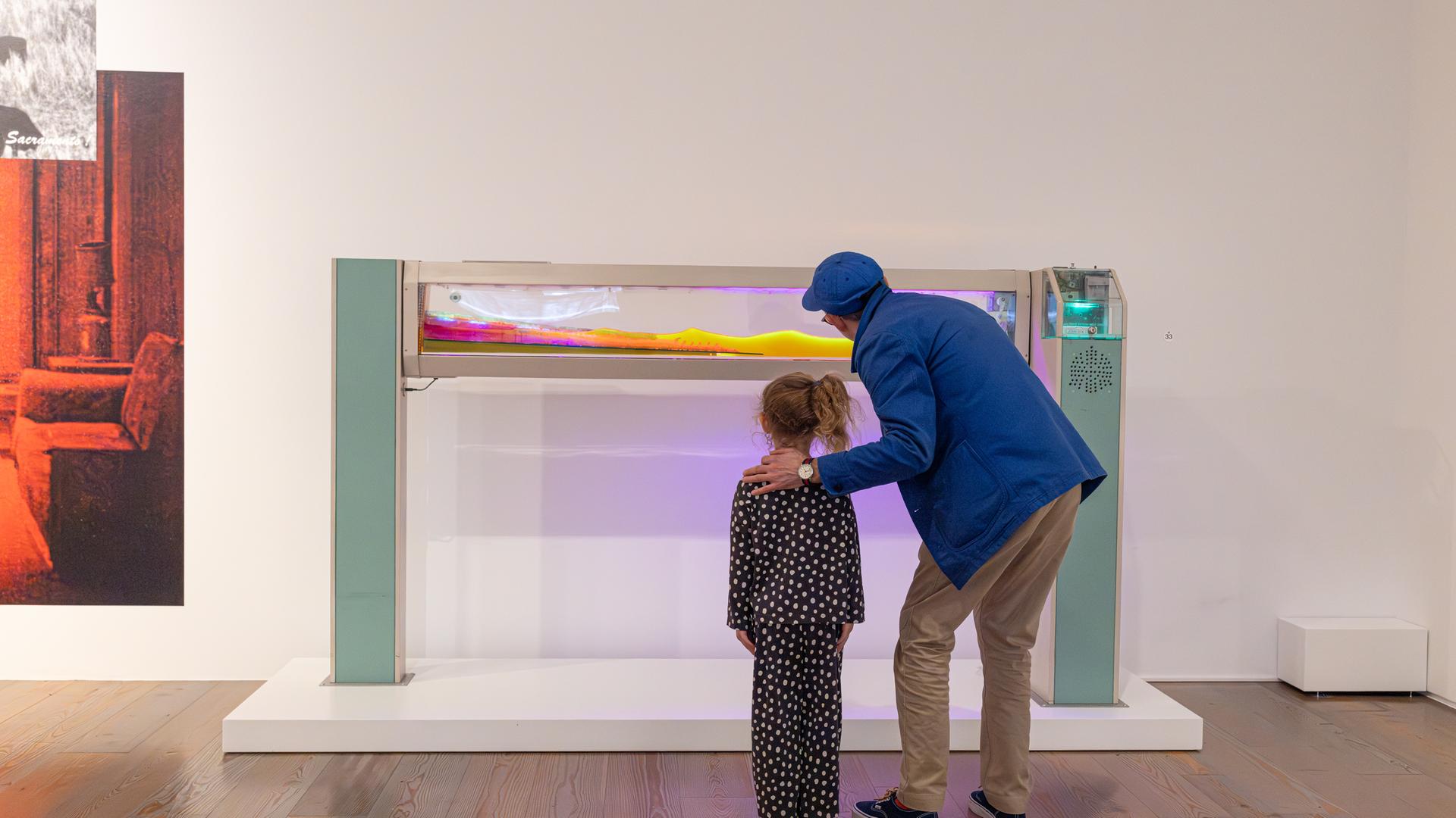 In einem Ausstellungsraum betrachten ein Mann und ein Mädchen eine Installation aus einem Glaskasten, in dem eine bunte Masse zu erkennen ist.