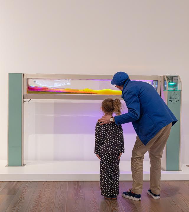 In einem Ausstellungsraum betrachten ein Mann und ein Mädchen eine Installation aus einem Glaskasten, in dem eine bunte Masse zu erkennen ist.