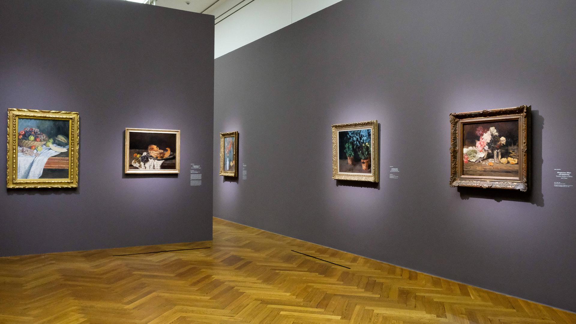 Zwei Wände an denen jeweils mehrere Gemälde hängen. Auf ein paar der Gemälde sind Blumen und Pflanzen zu sehen und auf einem ist eine Obstschale gemalt.