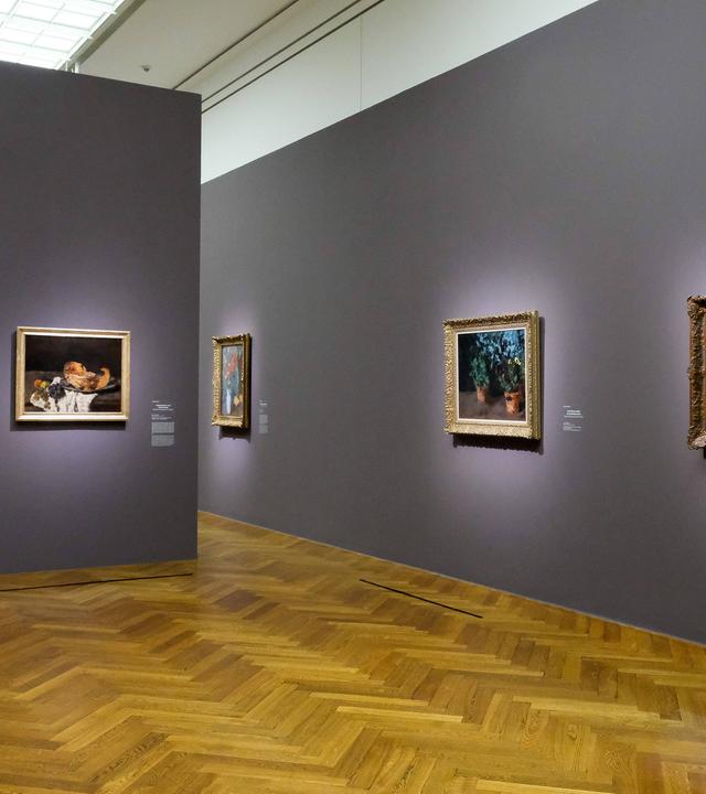 Zwei Wände an denen jeweils mehrere Gemälde hängen. Auf ein paar der Gemälde sind Blumen und Pflanzen zu sehen und auf einem ist eine Obstschale gemalt.