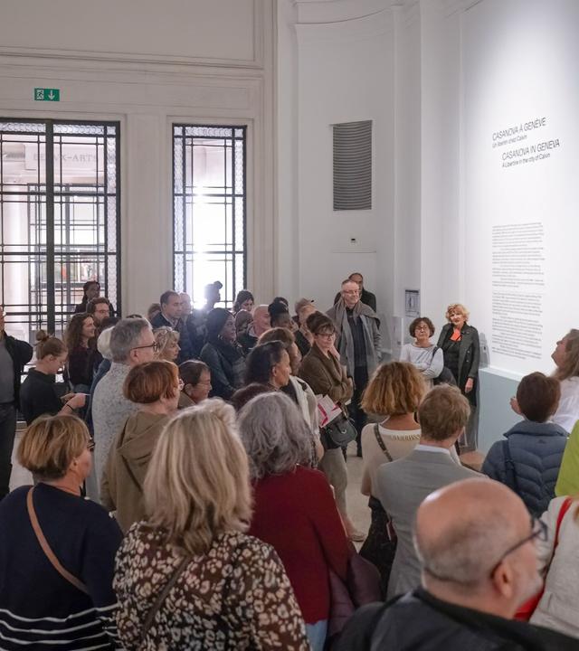 In einem Ausstellungsraum schauen sich viele Personen ein altes Gemälde rechts an der Wand an.