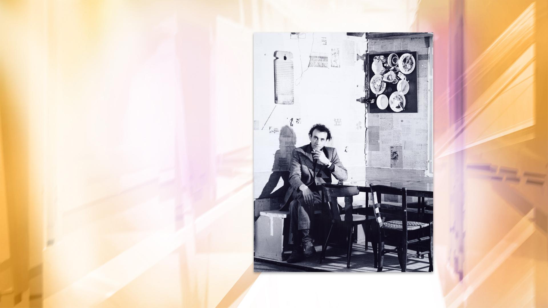 Auf einer Schwarz-Weiß-Fotografie sitzt ein Mann auf einer Bank in einer Ecke. An den Wänden hängt Geschirr.