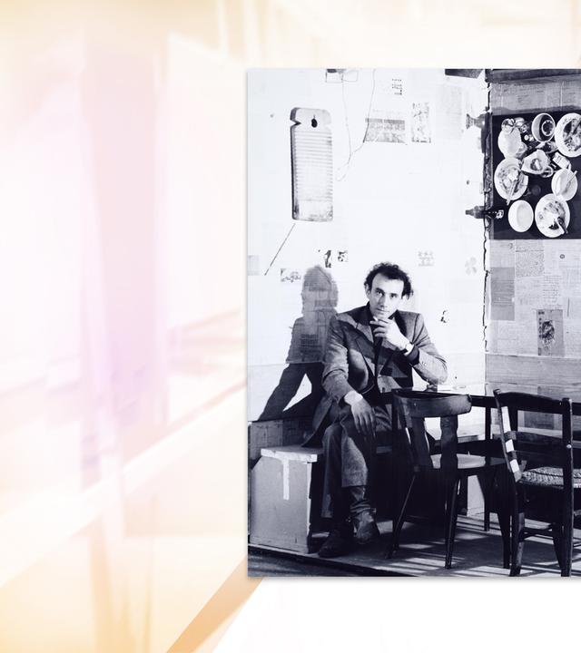 Auf einer Schwarz-Weiß-Fotografie sitzt ein Mann auf einer Bank in einer Ecke. An den Wänden hängt Geschirr.