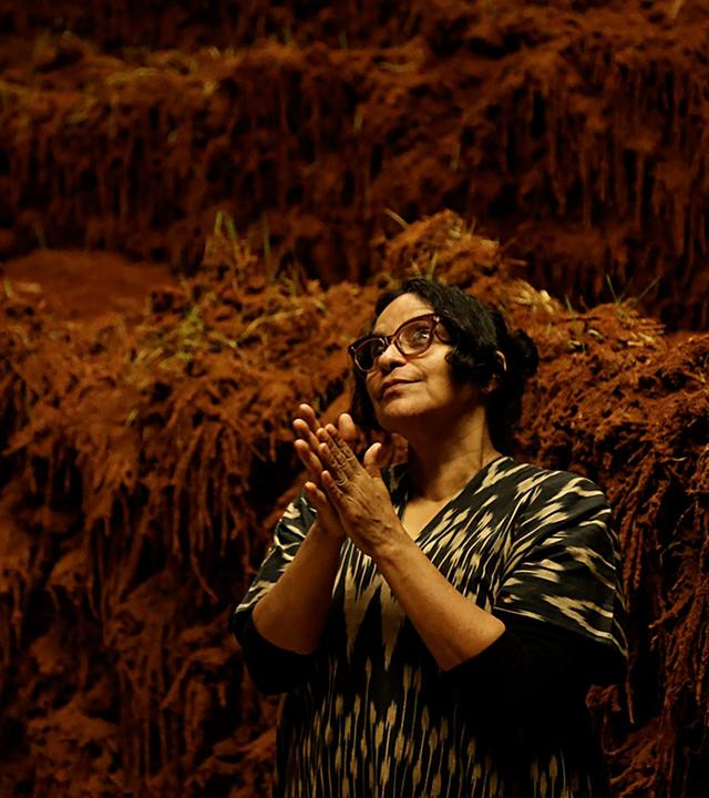 Man sieht eine Frau, die eine Brille trägt, vor einem Kunstwerk aus Erde.