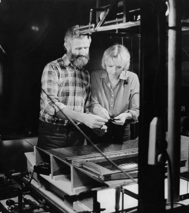 Auf der schwarz-weiß Fotografie sind ein Mann und eine Frau zu sehen, die vor einer Maschine stehen und auf etwas schauen, das sie in ihren Händen halten.