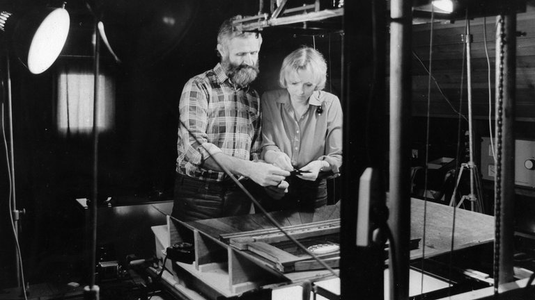 Auf der schwarz-weiß Fotografie sind ein Mann und eine Frau zu sehen, die vor einer Maschine stehen und auf etwas schauen, das sie in ihren Händen halten.