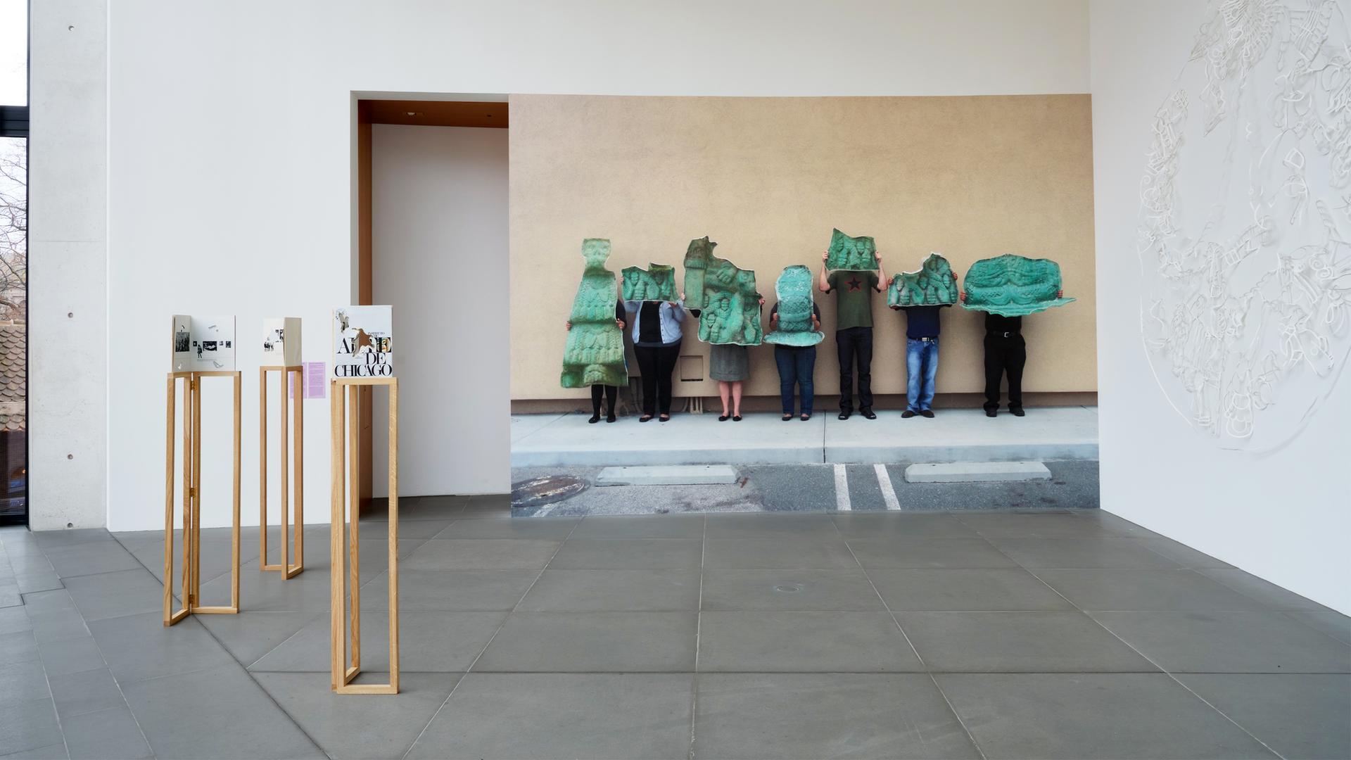 In einem Austellungsraum stehen Exponate und an der Wand befindet sich eine große Fotografe, die Menschen zeigt, die grüne Gegenstände vor sich halten.
