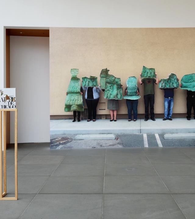 In einem Austellungsraum stehen Exponate und an der Wand befindet sich eine große Fotografe, die Menschen zeigt, die grüne Gegenstände vor sich halten.