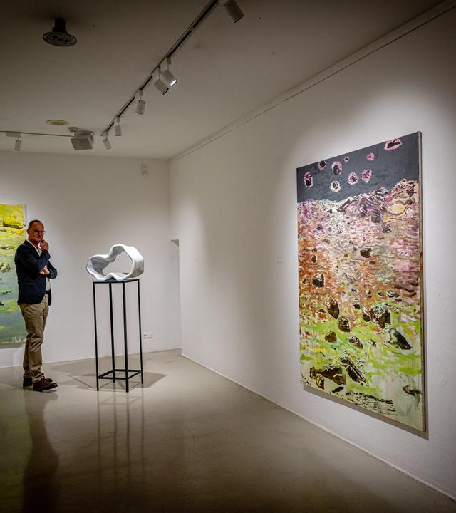 Ein Mann sieht sich eine moderne Skulptur in einem Ausstellungsraum an. An den Wänden hängen abstrakte Gemälde.