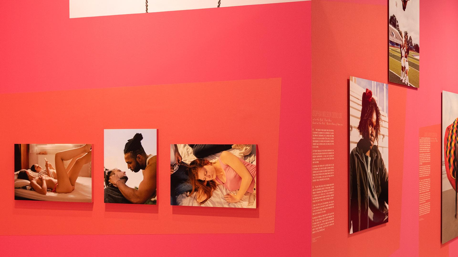 In einem Ausstellungsraum sieht man Fotografien von Menschen an einer roten Wand.