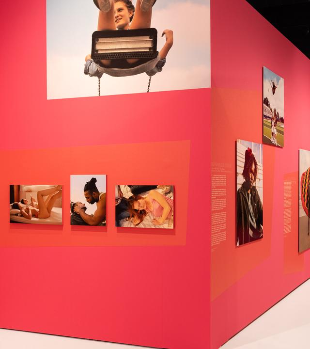 In einem Ausstellungsraum sieht man Fotografien von Menschen an einer roten Wand.