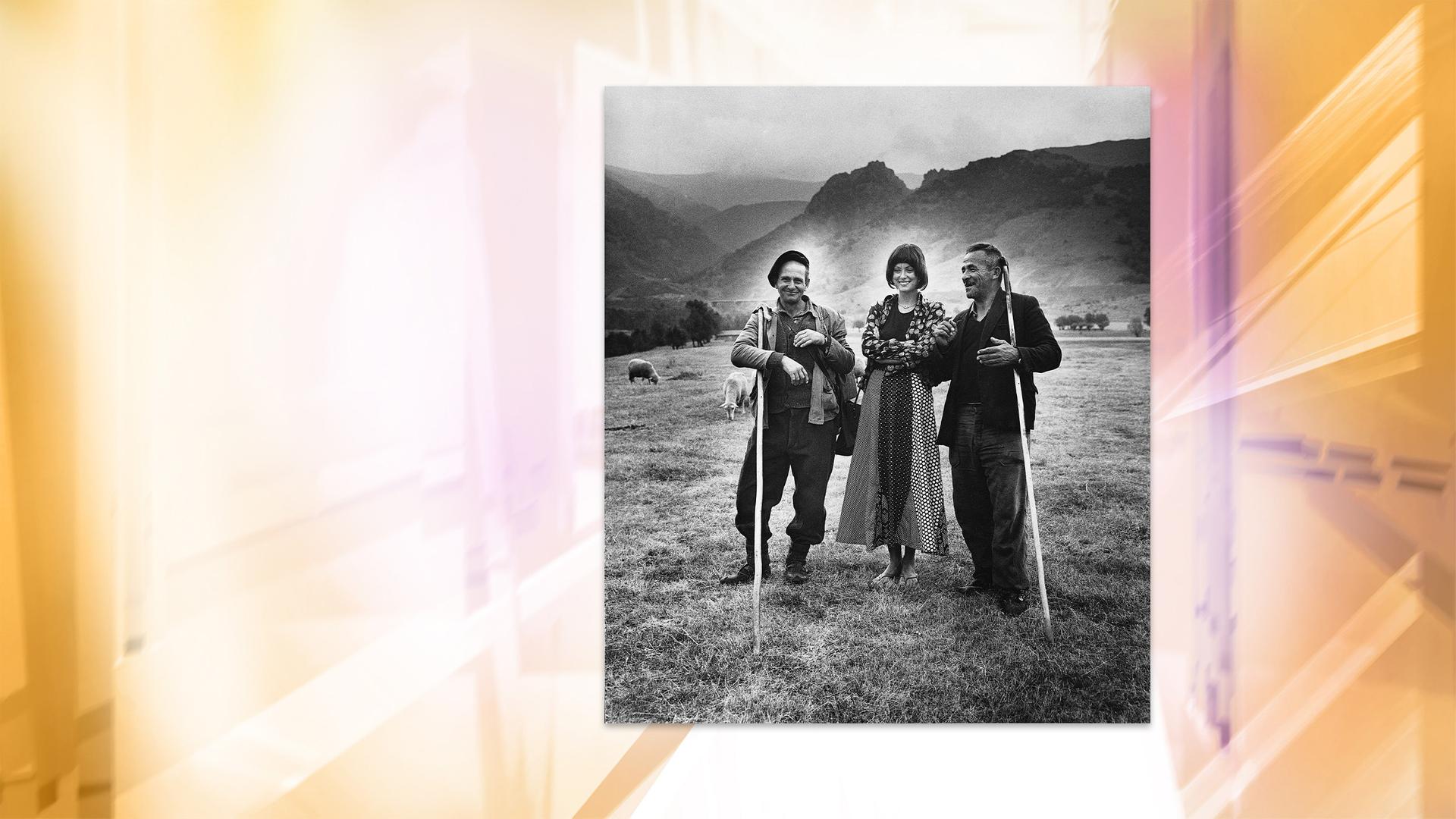 Auf einer Schwarz-Weiß-Fotografie sieht man eine Frau, die zwischen zwei Schäfern mit langen Stöcken auf einer Wiese vor hügeliger Landschaft steht.