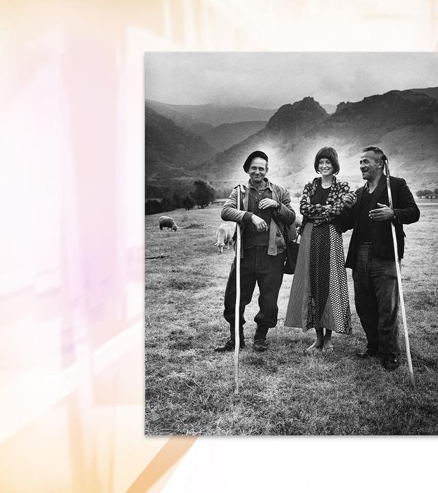 Auf einer Schwarz-Weiß-Fotografie sieht man eine Frau, die zwischen zwei Schäfern mit langen Stöcken auf einer Wiese vor hügeliger Landschaft steht.