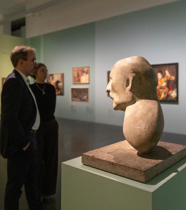 In einem Ausstellungsraum betrachten Menschen an der Wand hängende bunte Gemälde. Im Vordergrund steht die Büste eines Mannes.