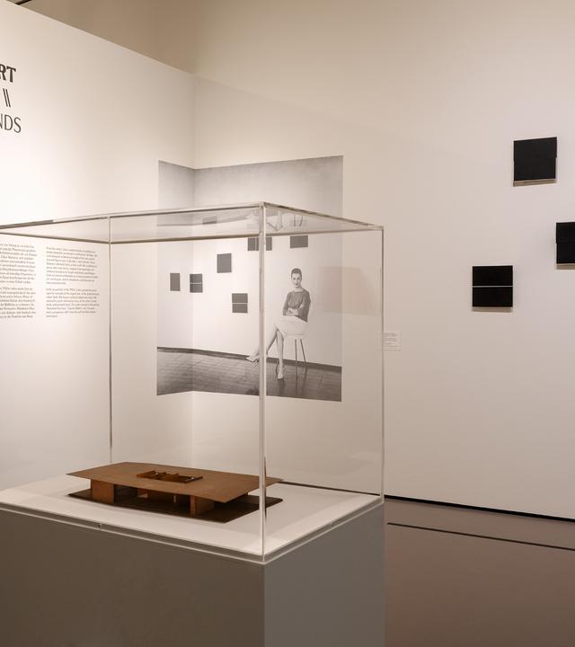 In einem Ausstellungsraum steht ein Glaskasten mit einem Exponat. An der Wand hängen Bilder und man sieht eine Schrift an der Wand.