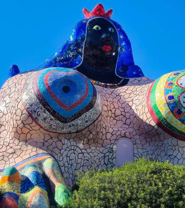 Man sieht ein Gebäude in Form einer voluminösen Frau, das mit buntem Mosaik geschmückt ist.