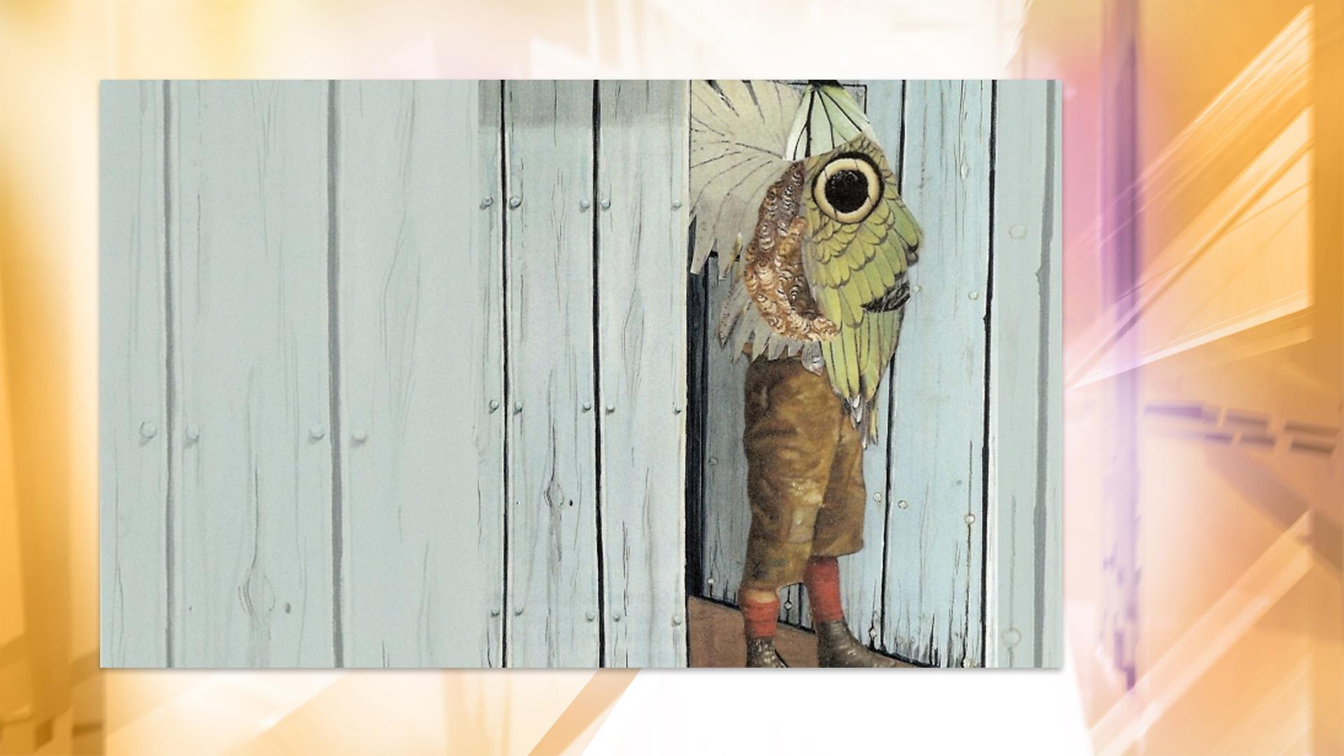 Man sieht eine Person in Lederhosen, die vor einer Holzwand steht. Sie trägt eine Art großen Schmetterlingsflügel der ihren Oberkörper bedeckt.