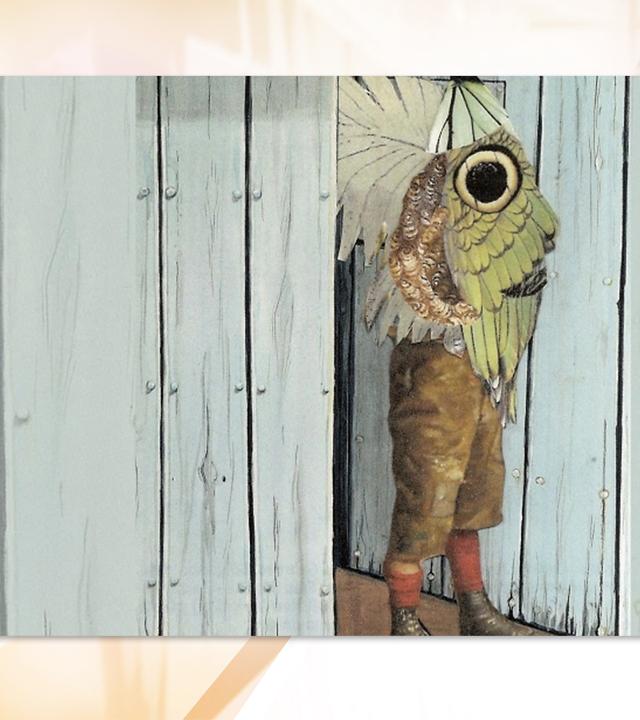 Man sieht eine Person in Lederhosen, die vor einer Holzwand steht. Sie trägt eine Art großen Schmetterlingsflügel der ihren Oberkörper bedeckt.