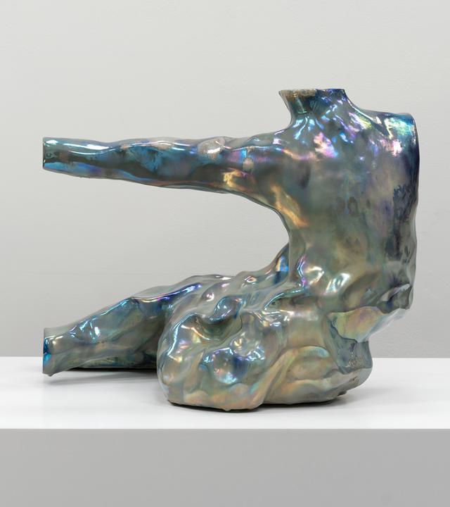 In einem Ausstellungsraum sieht man eine abstrakte, schillernde Figur, die an einen Menschen erinnert.