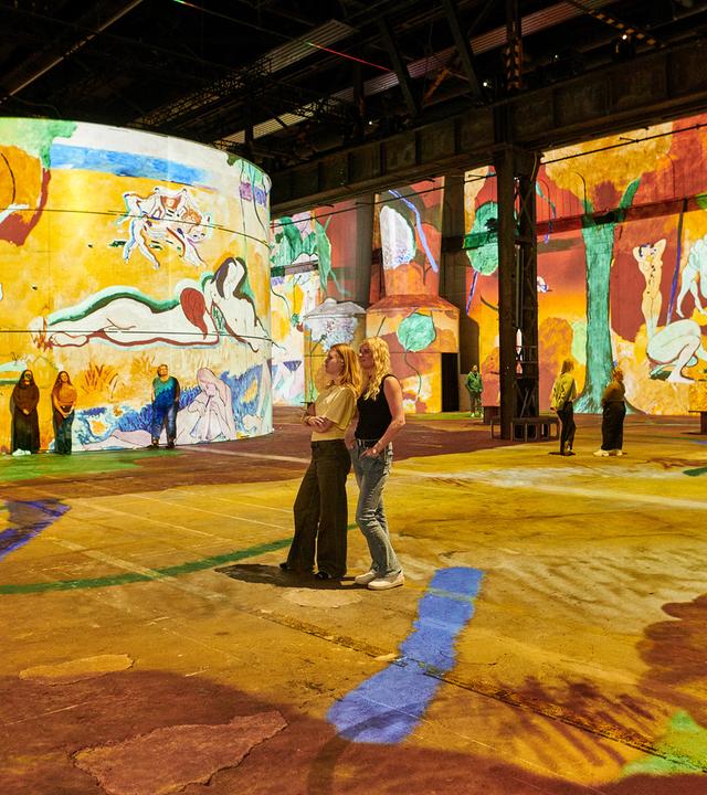 Menschen stehen und sitzen in einer großen Halle, an deren Wände und Boden bunte Gemälde projiziert werden.