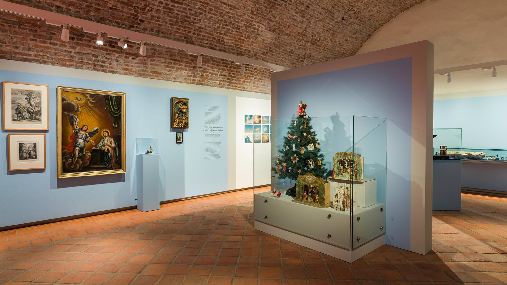 Zu sehen ist ein Raum der Ausstellung. Links an der Wand hängen mehrere Bilder, auf denen Engel zu erkennen sind. Rechts steht ein kleiner geschmückter Weihachtsbaum in einer Vitrine vor einer Wand.