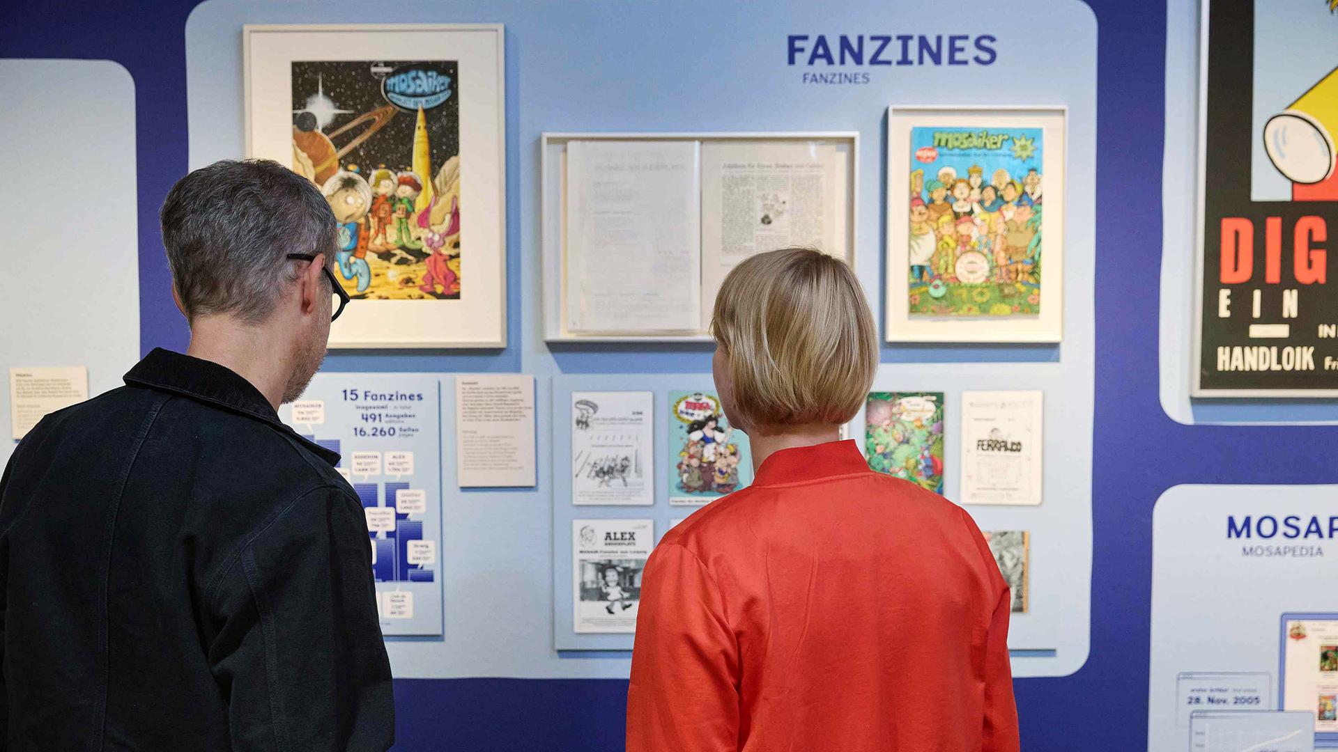 In einem Austellungsraum schauen sich ein Mann und eine Frau Comic-Exponate an.