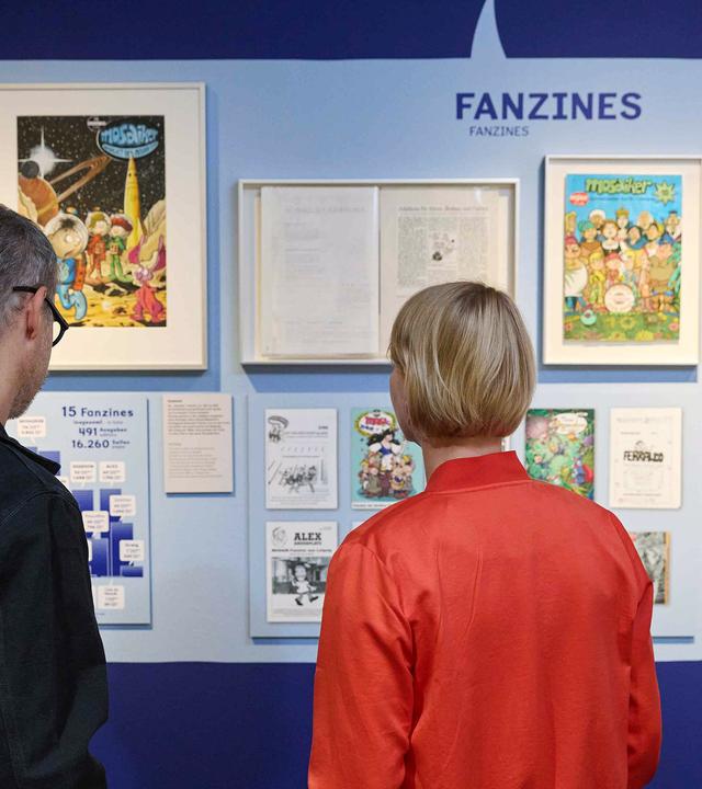 In einem Austellungsraum schauen sich ein Mann und eine Frau Comic-Exponate an.