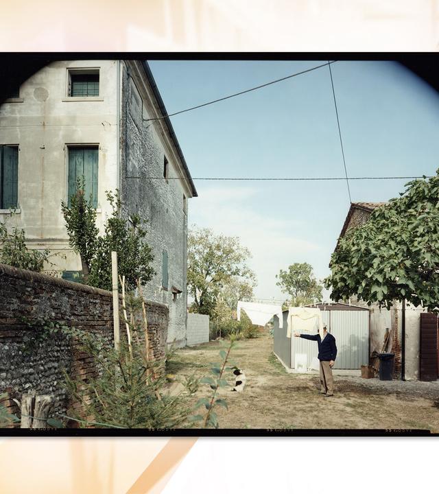 Ein Mann steht in einem Garten zwischen zwei Häusern. Vor ihm sitzt ein Hund, dem er ein Zeichen zu geben scheint. Das Gesicht des Mannes ist von einem Ärmel verdeckt, weil eine Wäscheleine durch den Garten gespannt ist, an der zwei Oberteile hängen. Im Garten stehen Bäume.