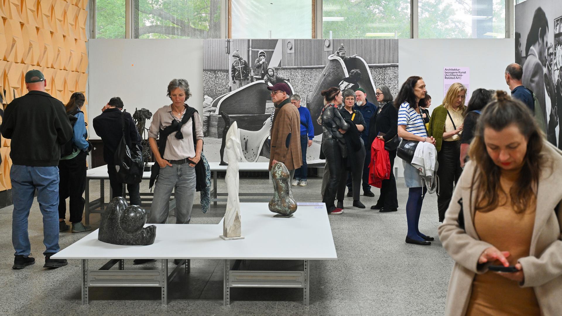 In einem Ausstellungsraum schauen Menschen sich die Plastiken, die auf Tischen stehen und großformatige Schwarz-weiß-Fotografien an den Wänden an.