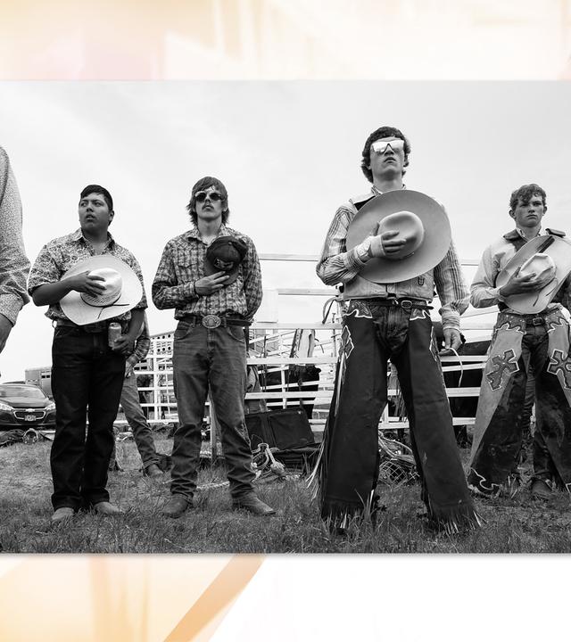 Auf einer Schwarz-Weiß-Fotografie sieht man mehrere breitbeinig nebeneinander stehende Cowboys , die sich jeweils ihren Hut auf die Brust halten.