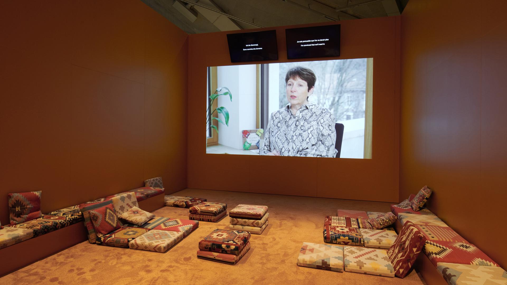 In einem Ausstellungsraum ist ein Monitor an der Wand, auf dem eine Frau zu sehen ist. Im vorderen Bereich stehen Sofas.