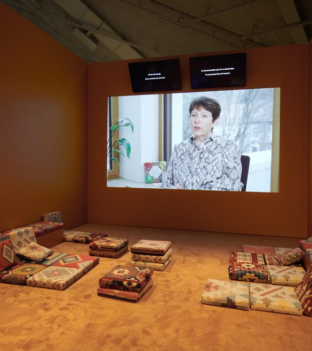 In einem Ausstellungsraum ist ein Monitor an der Wand, auf dem eine Frau zu sehen ist. Im vorderen Bereich stehen Sofas.