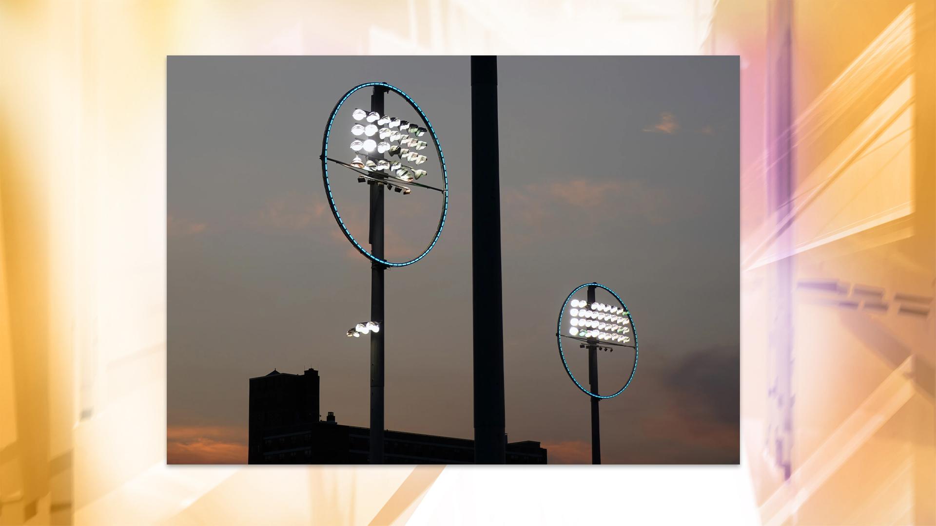 Eine Lichtinstallation, bestehend aus zwei hohen Stangen über einem Industriegebäude, auf denen je eine rechteckige Gruppe Strahler angeordnet ist, die von einem blau leuchtenden Ring umgeben ist.