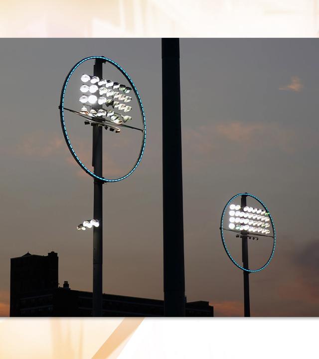 Eine Lichtinstallation, bestehend aus zwei hohen Stangen über einem Industriegebäude, auf denen je eine rechteckige Gruppe Strahler angeordnet ist, die von einem blau leuchtenden Ring umgeben ist.