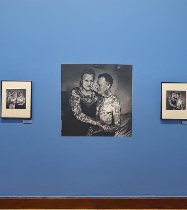 In einem Ausstellungsraum sieht man Schwarz-Weiß-Fotografien an einer blauen Wand. Die Fotografien zeigen tätowierte Menschen.