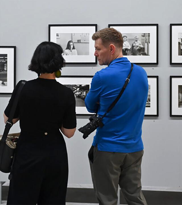 In einem Ausstellungsraum schauen eine Frau und ein Mann sich Schwarz-Weiß-Fotografien an einer Wand an.