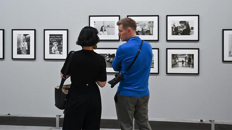 In einem Ausstellungsraum schauen eine Frau und ein Mann sich Schwarz-Weiß-Fotografien an einer Wand an.